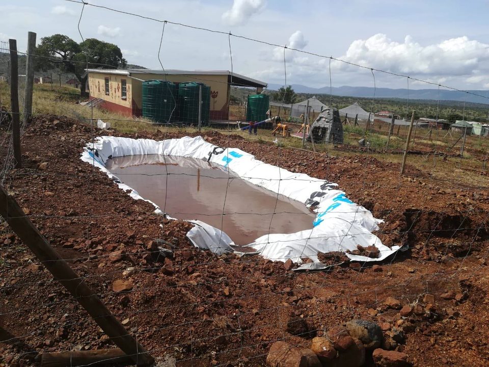 A newly lined holding pond filled with rainwater at Mzomoyethu Crèche, with tanks and growing structures beyond